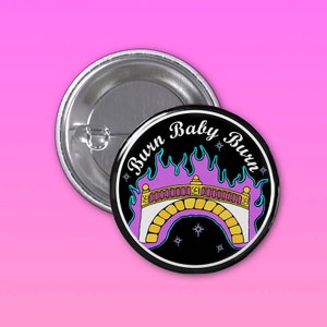 Puede incluir: Un botón redondo con fondo negro y la frase "Burn Baby Burn" en un círculo. El diseño muestra un puente con llamas moradas y detalles dorados. Se ve un segundo botón metálico sin diseño.