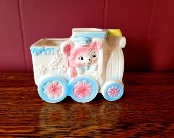 May include: A white ceramic train planter with a pink bear face on the front. The train has blue and pink wheels and is decorated with white flowers.