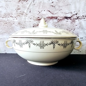 May include: A white ceramic serving bowl with a lid. The bowl has a black and gold floral design around the rim. The lid has a knob on top.