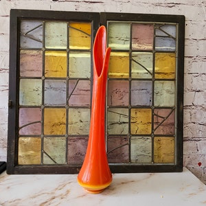 May include: A tall, orange glass vase with a flared opening and a yellow base. The vase is sitting on a white surface in front of a stained glass window.