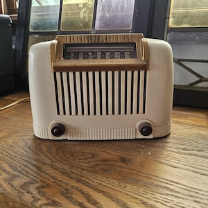 May include: Cream-coloured vintage Bendix radio with a gold-toned top and dial. The radio has a series of vertical black lines and two dark brown knobs. The radio sits on a wooden surface.