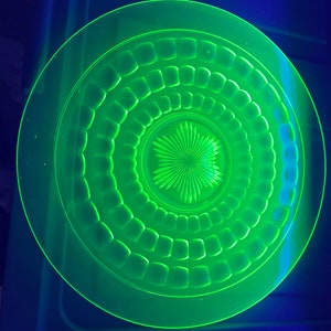 May include: A glowing green glass plate with a circular design. The plate features concentric rings of square and starburst patterns. The plate is illuminated with a bright green light, creating a vibrant and eye-catching effect.