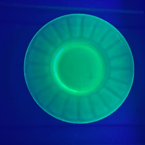 May include: A green glass plate with a fluted edge and a smaller plate in the center. The plate is illuminated with a black light, causing it to glow.