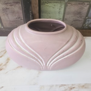 Puede incluir: Un jarrón de cerámica rosa claro con forma ovalada. Presenta detalles blancos curvos y un interior oscuro. El jarrón está sobre una superficie de mármol blanco. Aproximadamente 25 cm de ancho.