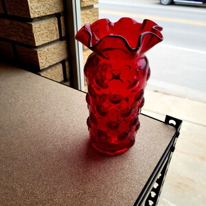 Könnte beinhalten: Eine rote Glasvase mit einem gekräuselten Rand und einem strukturierten, blasenartigen Design. Die Vase steht auf einer braunen Oberfläche.