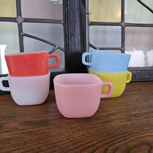 以下が含まれることがあります： 赤、白、ピンク、青、黄色のカラフルなヴィンテージスタイルのティーカップのコレクション。カップは積み重ねられ、木製の表面に配置され、背景には窓があります。