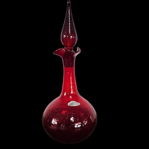 Może przedstawiać: Rubinowa szklana karafka z długim, spiczastym korkiem. Karafka ma pękatą podstawę i smukłą szyjkę. Szkło ma teksturowany, spękany wygląd. Do karafki przymocowana jest mała etykieta.