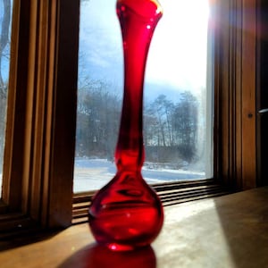 May include: A tall, red glass vase with a bulbous base. The vase is sitting on a wooden surface in front of a window.