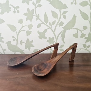 Puede incluir: Dos cucharas de madera con mangos únicos. Los mangos tienen forma de arco curvo y están hechos de madera marrón oscura. Las cucharas descansan sobre una superficie de madera.
