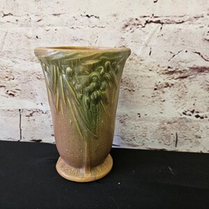 Puede incluir: Un jarrón de cerámica marrón y verde con un diseño floral. El jarrón tiene una boca ancha y una base estrecha.
