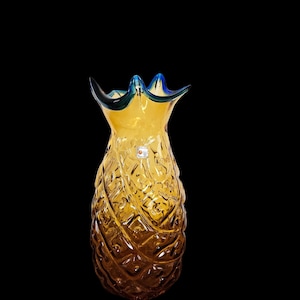 May include: A decorative glass vase shaped like a pineapple. The vase is amber-colored with a textured surface and a blue and green rim. The pineapple design is detailed and the vase is set against a black background.