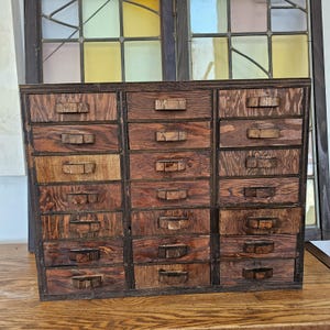 Può includere: Un mobiletto in legno marrone scuro con ventuno cassetti singoli disposti in tre colonne verticali. Ogni cassetto è dotato di una semplice maniglia rettangolare in legno centrata sul pannello frontale, che mette in risalto le venature del legno.