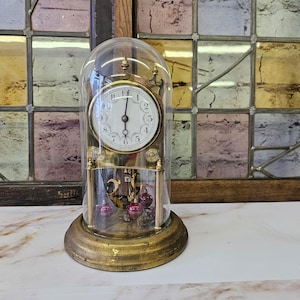 May include: An antique brass-toned clock with a glass dome. The clock face is white with black numerals and hands. The base has a circular design, and the pendulum features three red spheres. The clock is approximately 25 cm tall.