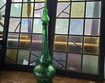 May include: A tall, green glass bottle with a long, slender neck and a bulbous base. The bottle has a decorative, pointed stopper. The background features a multi-coloured stained glass window, adding a vintage aesthetic.