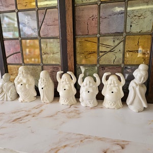 May include: Eight white ceramic figurines of the laughing Buddha, some with arms raised, and one with a traditional robe. The figurines are arranged on a marble surface.