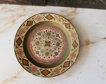 May include: A round brass plate with a decorative design in shades of pink, green, gold, and white. The design features intricate floral patterns and geometric shapes.
