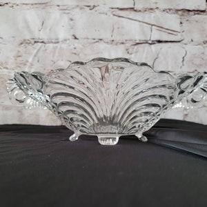 May include: A clear glass serving bowl with a shell design. The bowl has a scalloped rim, two handles, and three small feet. The bowl is approximately 10 inches wide and 4 inches tall.
