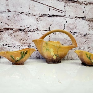 May include: A set of three vintage ceramic teacups in a yellow glaze with a floral design. The teapot has a unique curved handle and a spout. The cups are small and have a rounded shape.