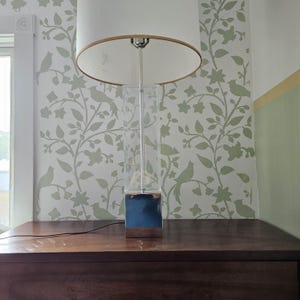 May include: A silver table lamp with a clear glass base and a white lampshade. The lamp is sitting on a wooden table.