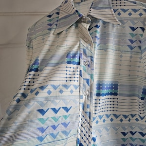 May include: A button-down shirt with a geometric pattern in shades of blue and white. The shirt features a collared neckline and is hanging on a black hanger. The pattern includes lines, diamonds, and triangles.