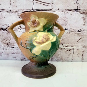 May include: A brown and green ceramic vase with two handles and a floral design featuring white magnolia flowers. The vase has a textured surface and a round base.