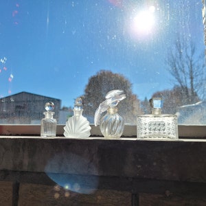 May include: Four clear glass perfume bottles with different shaped stoppers and designs. The bottles are arranged in a row on a windowsill. The background is a blurry view of a blue sky and trees.