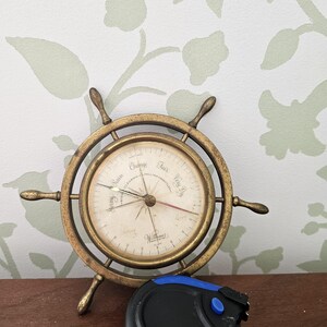 May include: A brass ship's wheel barometer with a white face and black markings. The barometer has a needle that points to the current weather conditions, including "stormy rain", "change", "fair", and "very dry". The barometer is made by the company "Wilh. Lammers".
