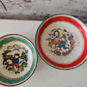 May include: Two small, round decorative plates with colorful folk art designs. One plate has a red border and depicts figures and flowers. The other has a green border and features a deer and a person.