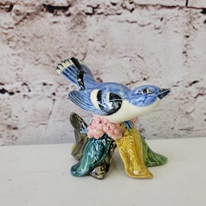 Puede incluir: Una figurita de pájaro de cerámica azul y blanca posada sobre una base floral colorida. El pájaro tiene un ojo negro y un pico amarillo. La base está decorada con flores rosas, verdes y amarillas.