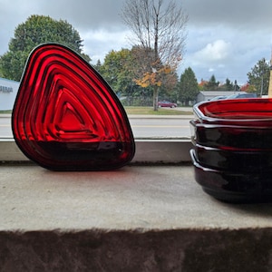 May include: Two red glass dishes, one shaped like a triangle and the other a square, are sitting on a windowsill. The dishes are made of ruby red glass and have a layered, textured design.