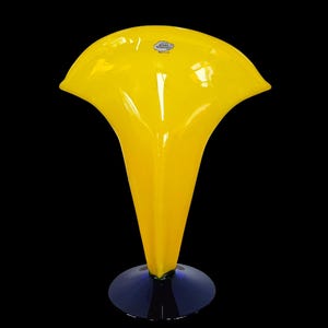 Può includere: Un vaso di vetro giallo brillante con una parte superiore svasata e un corpo a forma di tromba. Poggia su una base circolare blu scuro. Una piccola etichetta bianca è attaccata in alto. Il vaso è alto circa 30 cm.