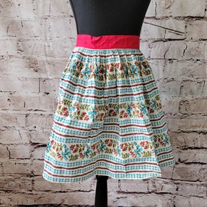 May include: A white apron with a blue and red floral pattern and a solid pink waistband. The apron is on a mannequin against a white brick wall.