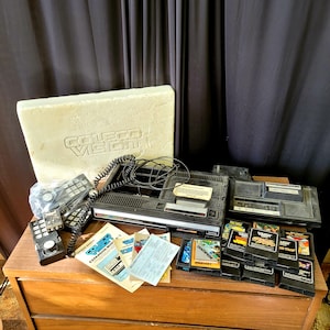 May include: A retro ColecoVision games console, controllers, game cartridges, and manuals. The console is black and grey, with a cream-coloured box. Game cartridges include titles such as "Centipede" and "Jungle Hunt".