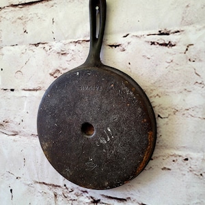 May include: A vintage cast iron skillet with a black handle. The skillet has a round shape and a hole in the center. The text "TIVOLIZE" is visible on the skillet.
