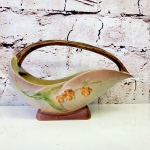 Puede incluir: Un jarrón de cerámica vintage con un asa marrón y un diseño floral. El jarrón es de color rosa claro, verde y amarillo con un asa marrón. El jarrón tiene forma de hoja y una base cuadrada.