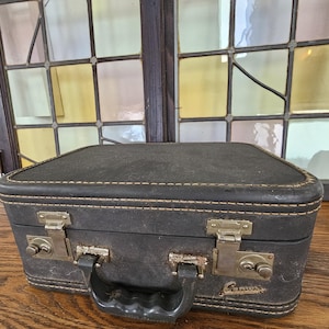 May include: A vintage black suitcase with a handle and two metal latches. The suitcase has a rectangular shape with rounded corners and is stitched with a light-colored thread. The metal latches and handle show signs of age.