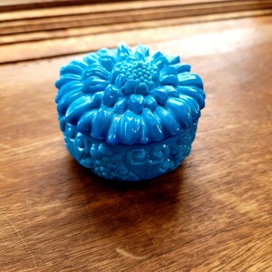 May include: A small, blue, ceramic trinket box with a floral design. The lid features a raised, stylized flower with petals and a center.