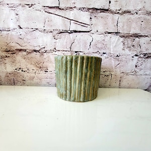 Op de afbeelding: Een groene keramische plantenpot met verticale lijnen. De pot heeft een lichtgroene glazuur met een subtiele gouden glans.