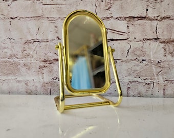May include: A gold-toned tabletop vanity mirror with a rounded top and a stand that can be adjusted to different angles.
