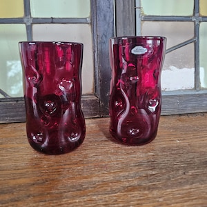 May include: Two ruby red drinking glasses with a textured, bubble-like design. The glasses are cylindrical with a slightly curved shape and sit on a wooden surface. The background includes a window with stained glass.