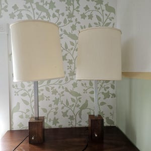 May include: Two white lampshades with silver bases and wooden blocks on a dark wood surface. The lampshades are cylindrical and have a slightly textured fabric.