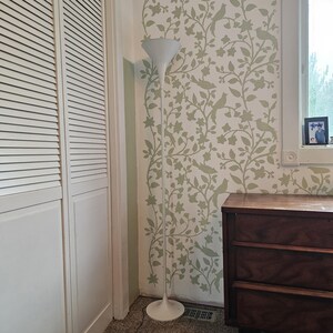 Peut inclure: Un lampadaire blanc avec un abat-jour blanc et un pied blanc. La lampe est placée dans une pièce aux murs blancs et une porte blanche. Le mur est recouvert d'un papier peint floral vert et blanc avec un motif d'oiseaux et de branches.