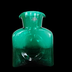 Può includere: Un vivace vaso di vetro verde smeraldo con un corpo unico e arrotondato e una parte superiore svasata e aperta. Il vaso fatto a mano ha un'etichetta vicino alla parte superiore. Il vetro trasparente lascia passare la luce, creando un gioco di ombre.