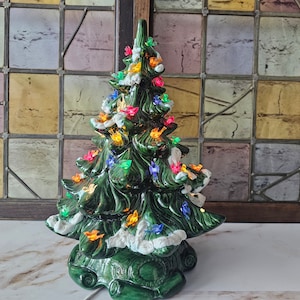 May include: A green ceramic Christmas tree with white snow and colorful lights. The tree is decorated with small birds and has a base with a scroll design.