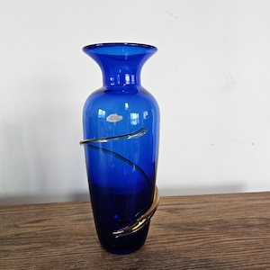 Può includere: Un vaso di vetro blu cobalto con bordo svasato e un accento dorato a spirale. Il vaso ha una finitura liscia e lucida ed è appoggiato su una superficie di legno. Un oggetto decorativo.