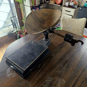 Puede incluir: Balanza antigua de color negro y latón con un cuenco redondo y una plataforma. La balanza está sobre una mesa de madera.