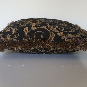 May include: A square cushion with a black background and a gold floral pattern. It has a brown fringe trim. The cushion is on a white surface. The cushion measures approximately 40cm x 40cm.