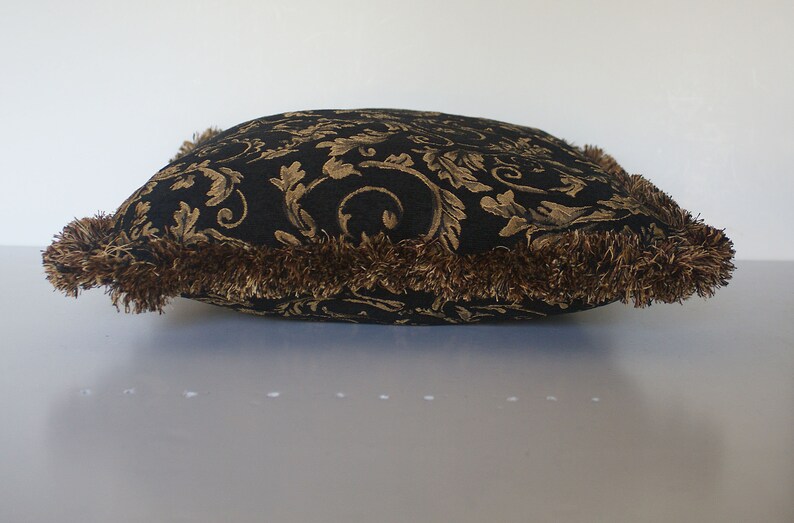 May include: A square cushion with a black background and a gold floral pattern. It has a brown fringe trim. The cushion is on a white surface. The cushion measures approximately 40cm x 40cm.