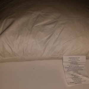 May include: A white pillow with a white fabric label attached to the bottom right corner. The label contains information about the pillow, including the manufacturer, care instructions, and size. The pillow is made of 100% cotton and is filled with down feathers.