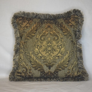 May include: A square throw pillow featuring a woven damask pattern in shades of olive green, gold, and dark brown. The pillow is finished with a thick, multi-toned fringe trim along all four edges, creating a textured border around the central design.